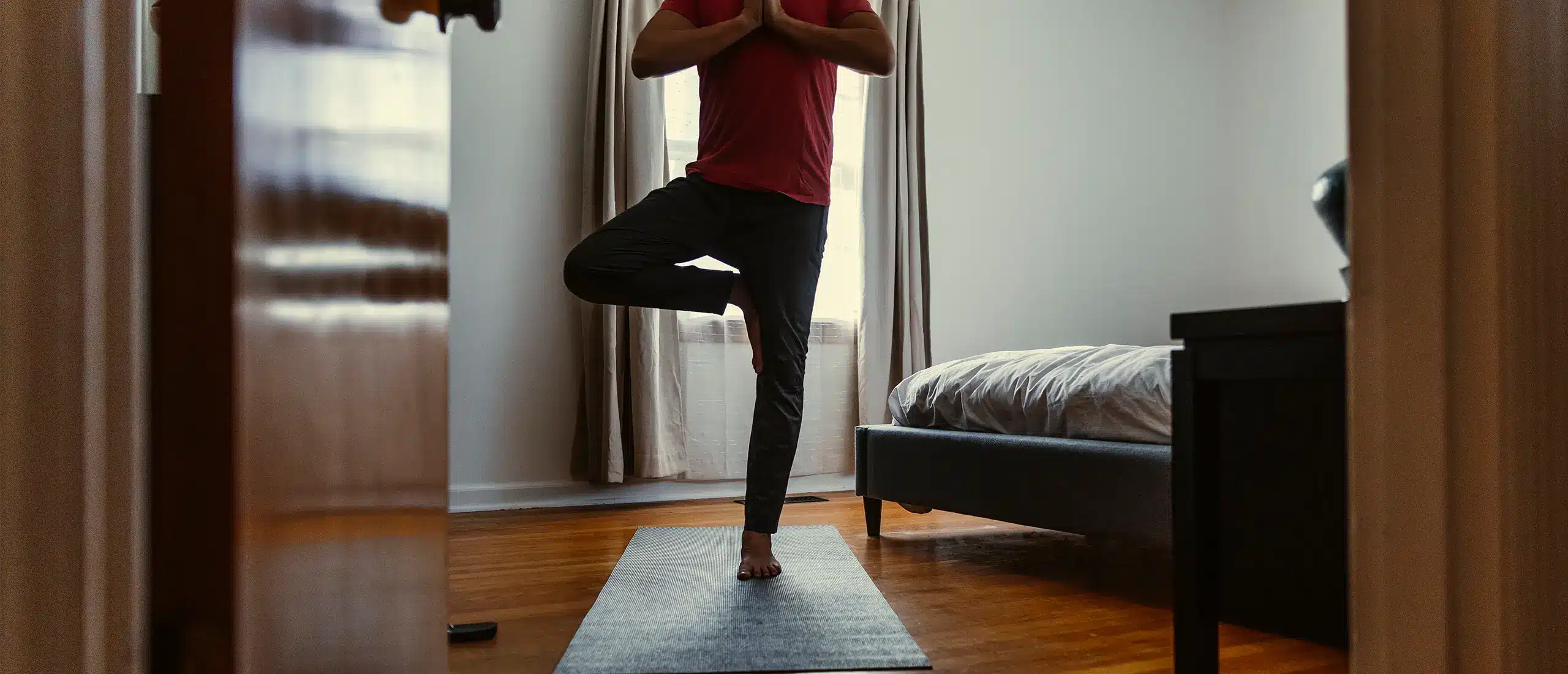 Man balancing on one foot on a yoga mat in a bedroom.