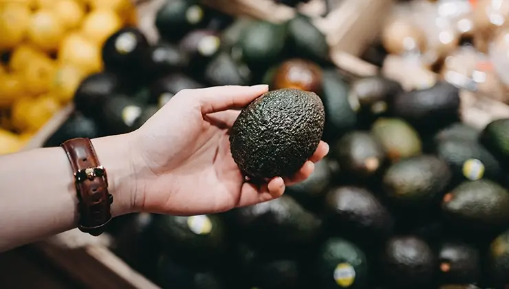 a woman plucks a ripe avocado out of a bin with multiple of them