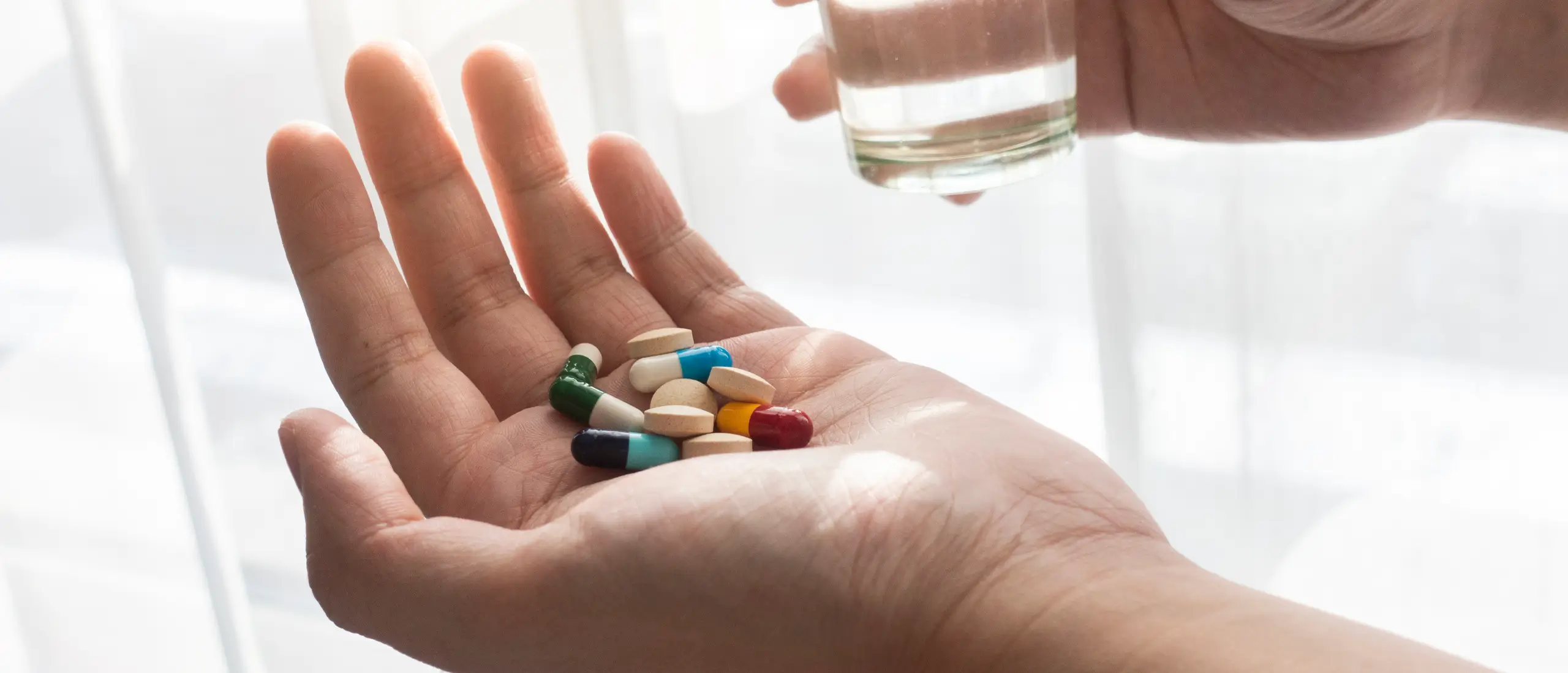 person holds out hand with an array of colorful pills. The other hand holds a glass of water
