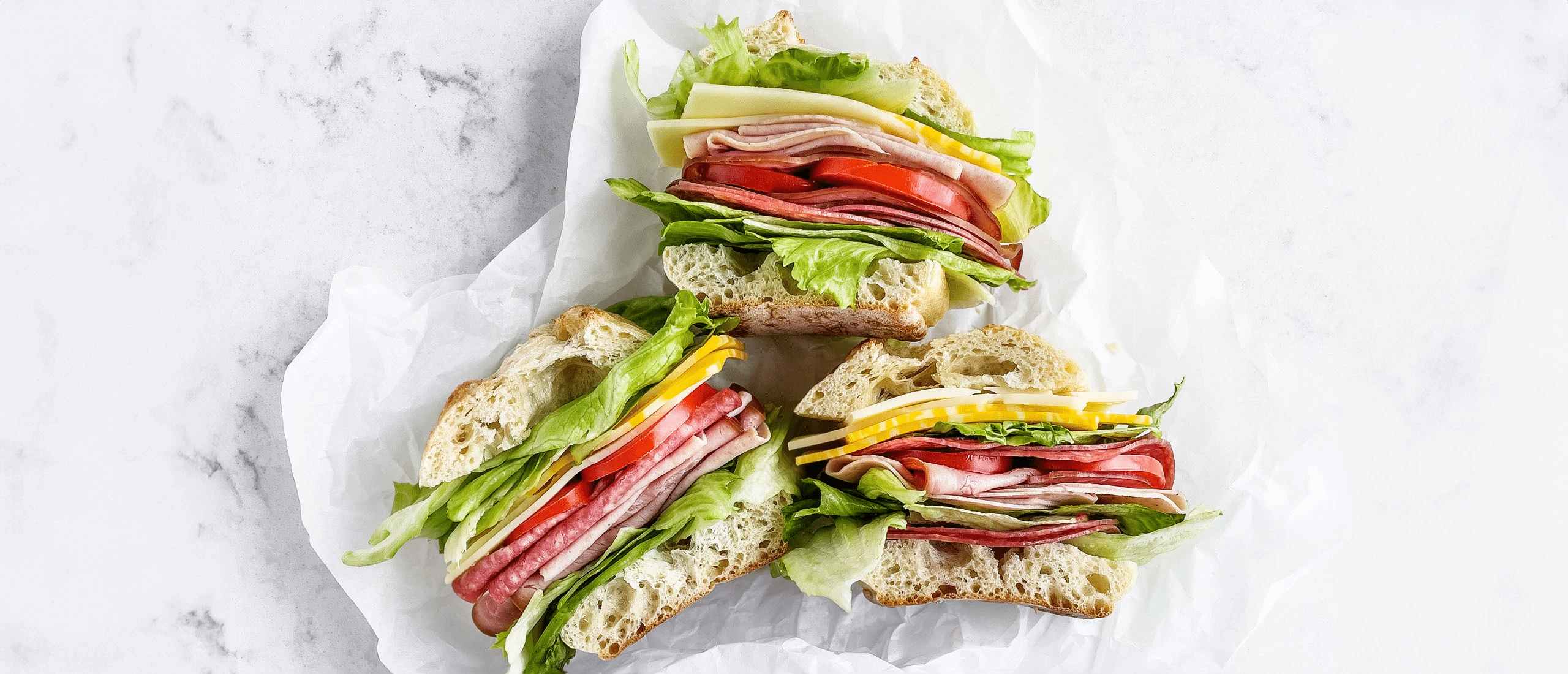 Three sandwiches loaded up with the works: deli meat, veggies, and cheese in wrappers on a white marble background